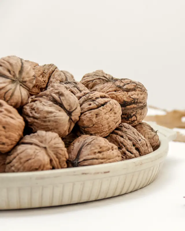 A shallow bowl of walnuts on a pale surface with dried oak leaves.